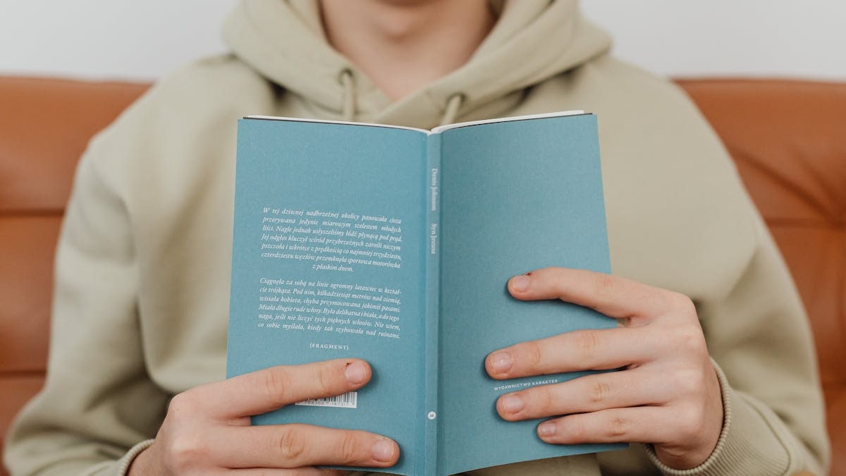 Close-up of hands holding a hardcover book, wearing a beige hoodie. Ideal for reading-related content.